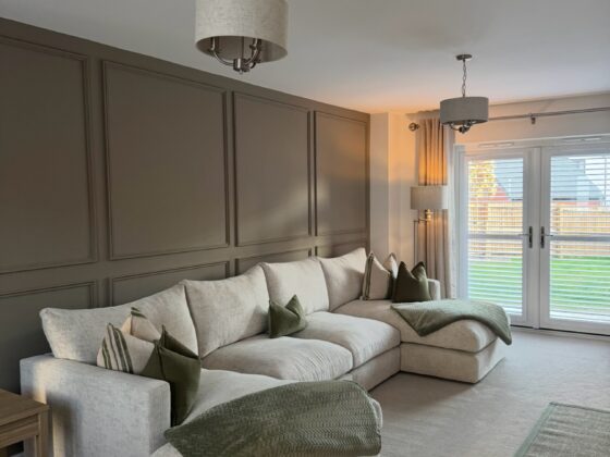 A sofa and side table-living room furniture-beige U-shaped corner chaise sofa-green scatter cushions-green throw-green edged jute rug-beige carpet-brown beaded feature wall-double doors with shutters-floor standing lamp-beige and metal ceiling lights-wooden side table-off white ceiling