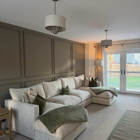 A sofa and side table-living room furniture-beige U-shaped corner chaise sofa-green scatter cushions-green throw-green edged jute rug-beige carpet-brown beaded feature wall-double doors with shutters-floor standing lamp-beige and metal ceiling lights-wooden side table-off white ceiling