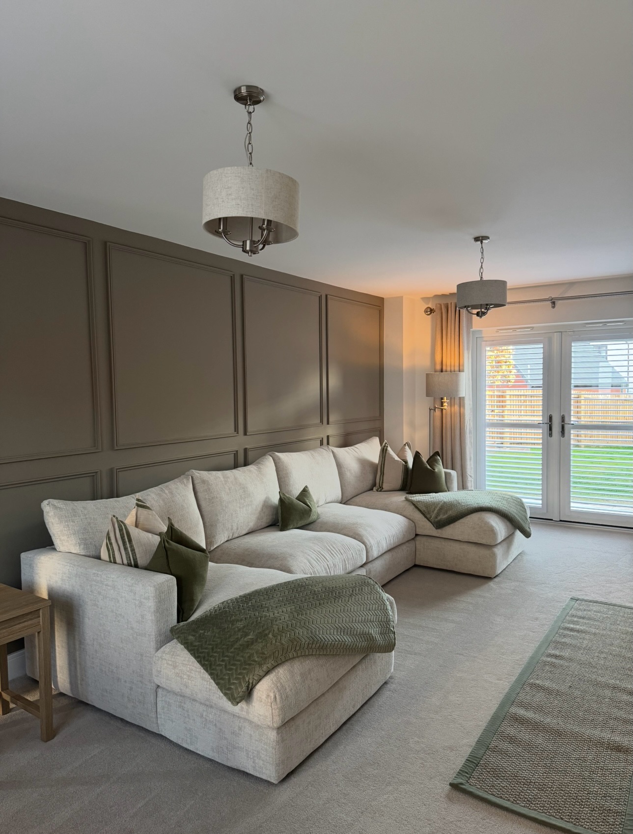 A sofa and side table-living room furniture-beige U-shaped corner chaise sofa-green scatter cushions-green throw-green edged jute rug-beige carpet-brown beaded feature wall-double doors with shutters-floor standing lamp-beige and metal ceiling lights-wooden side table-off white ceiling