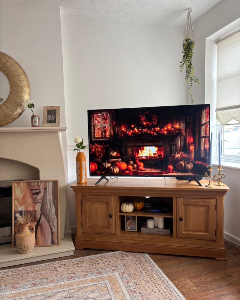 A TV unit-dining room furniture-wooden TV unit with storage-Halloween pumpkin scene on TV screen-wooden floor-vintage style rug-beige fireplace-round brass mirror-pumpkin decor-hanging plant-white walls-brass candleholders