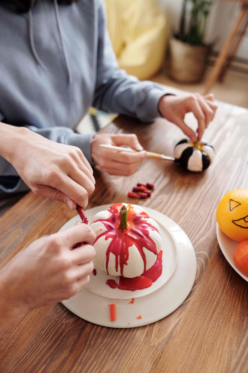 A table-dining room furniture-wooden tabletop-painting pumpkins-hands painting-paintbrushes-potted plant-decorated oranges in bowl