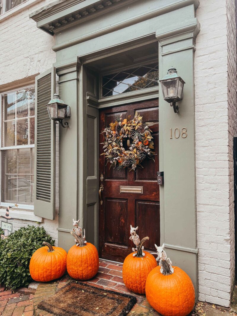A front door-house fascade-autumn wreath-pumpkins-skeleton decor-green entrance-dark brown door-window shutters-white painted brick