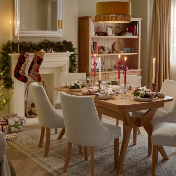 A dining table, chairs, bench, and dresser-dining room furniture-a wooden dining table-beige fabric dining chairs-large wooden dresser-wooden bench-draped curtains-marble patterned crackers-gold cutlery-christmas styling-white gravy boat-tapered candles in red and cream-delicate blue scalloped glassware-white crockery-off white fireplace-festive garland-christmas tree-handmade stockings-mustard tassel ceiling lampshade-wooden bench-vintage style rug-wooden parquet flooring